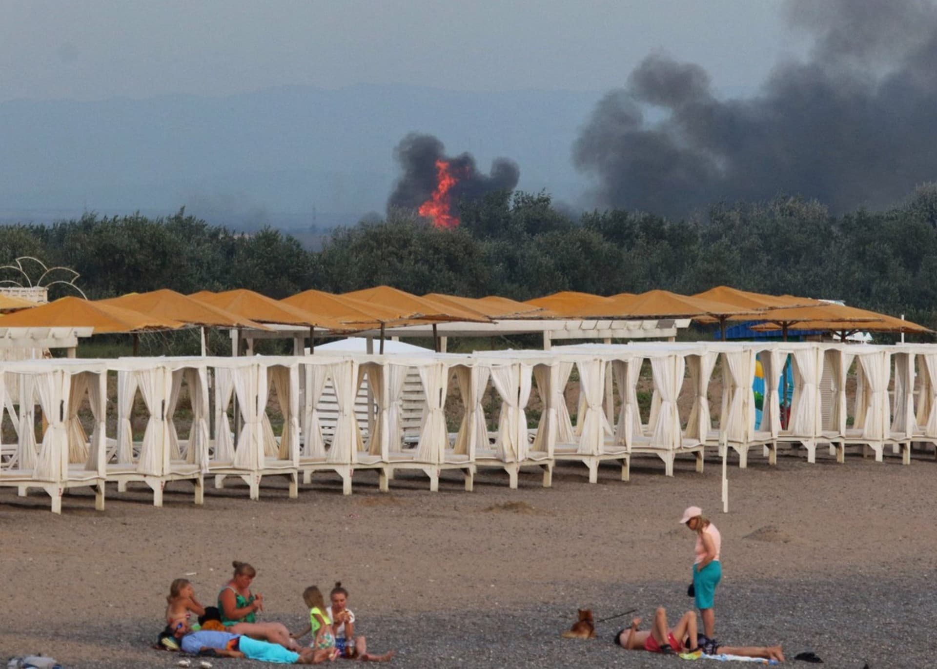 People rest on a beach as smoke and flames rise after a series of explosions at a Russian military airbase in Crimea