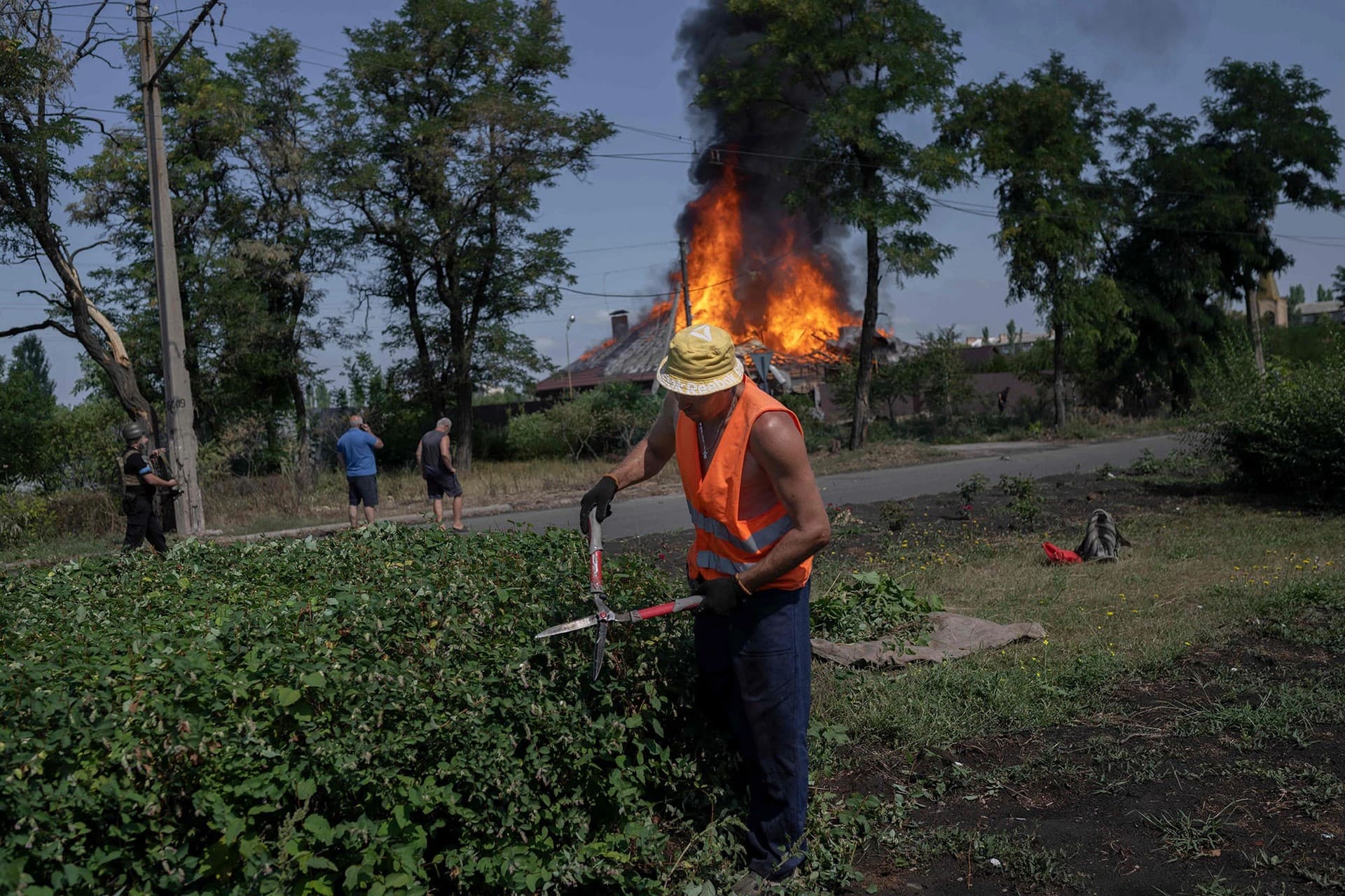 municipal worker trims hedges against the backdrop of a burning house in Bakhmut