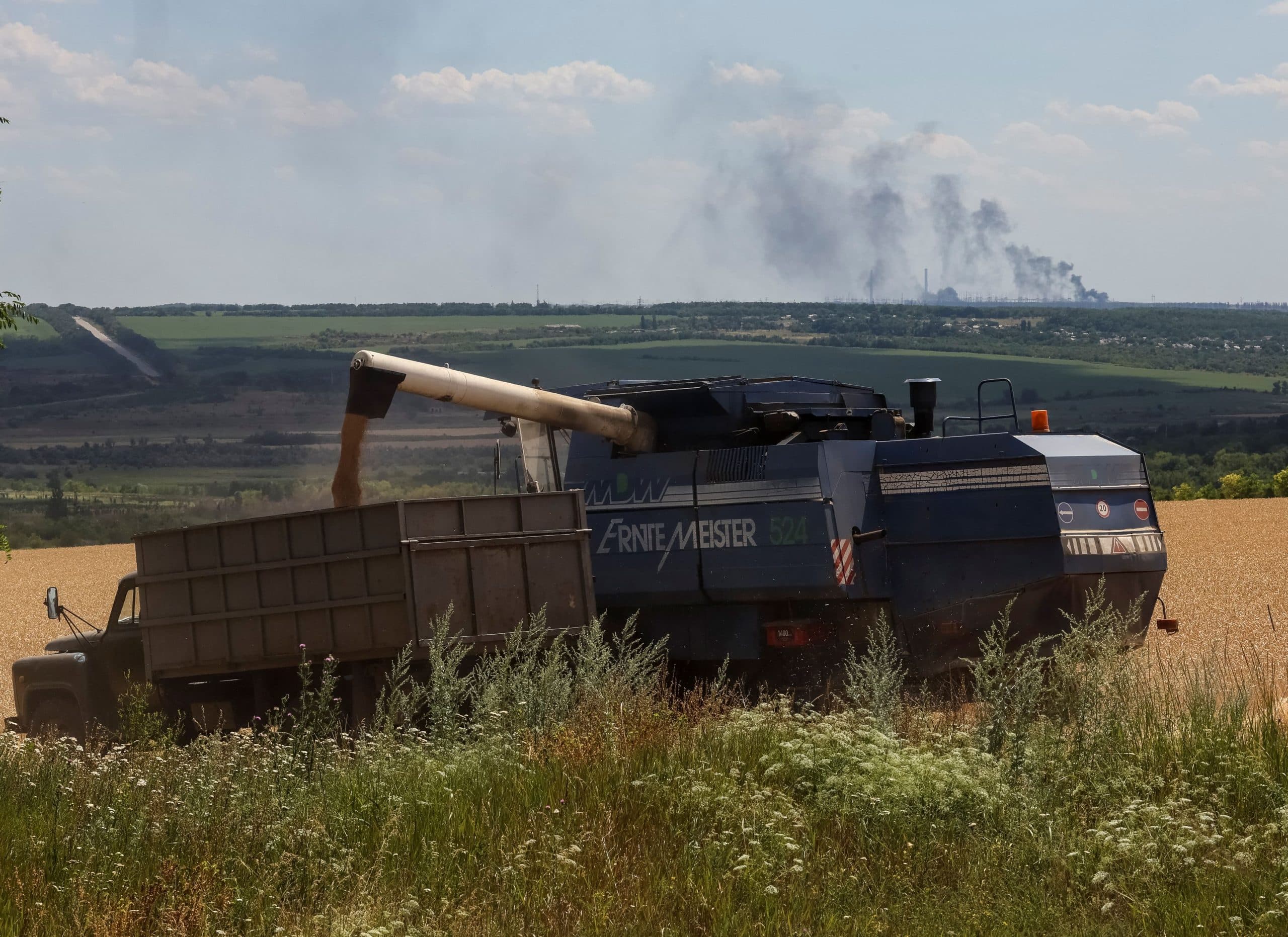 Farmers harvest wheat as Vuhlehirsk’s power plant burns in the distance after shelling