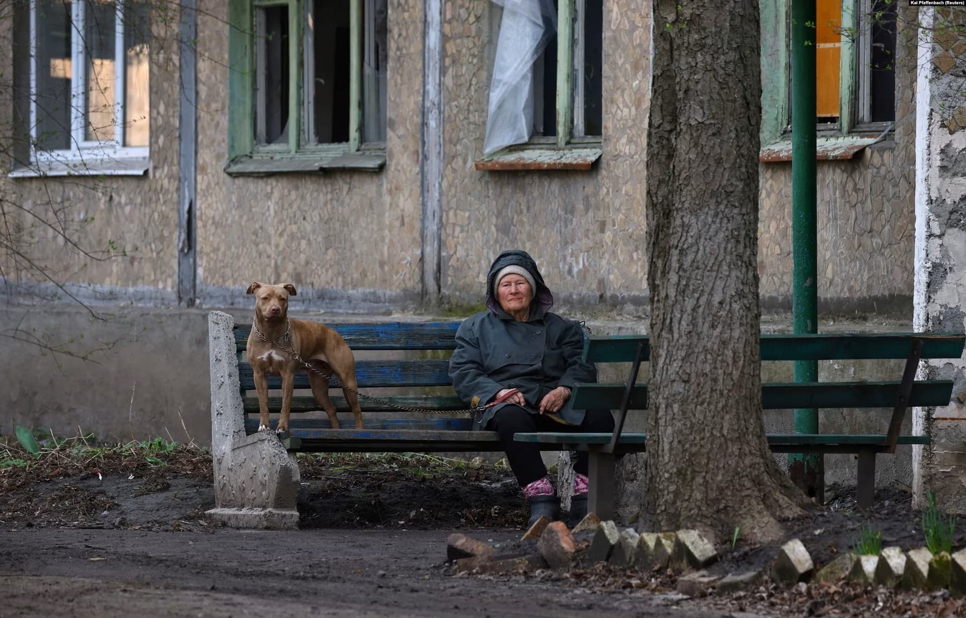 A woman sits with her dog on a bench outside a damaged apartment building in Chasiv Yar