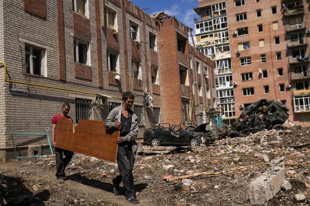 Two men carry a wooded panel next to heavily damaged buildings and destroyed cars in a Russian bombing in Bakhmut