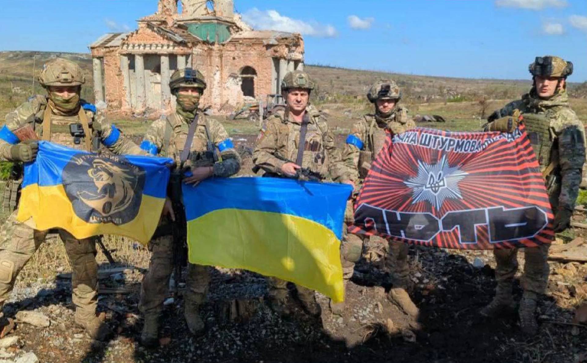 Ukrainian servicemen pose with Ukraine's national flag in front of a destroyed building in the village of Klishchiivka