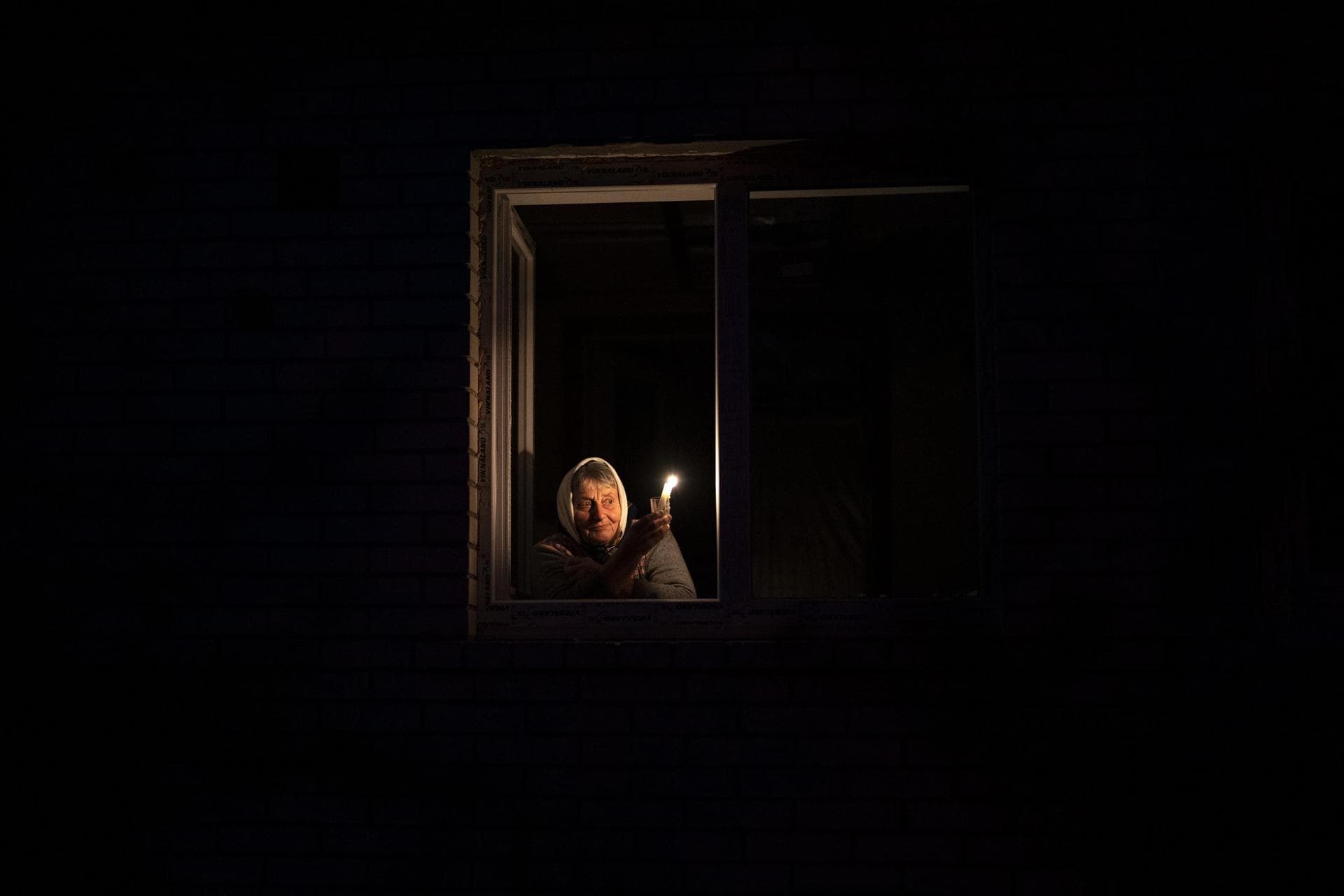 Catherine looks out the window while holding a candle for light inside her house during a power outage, in Borodyanka