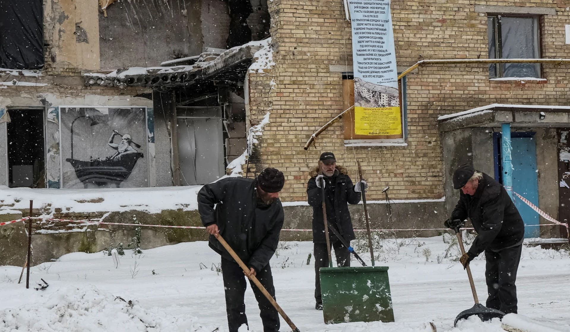 Local residents shovel snow as a work of world-renowned graffiti artist Banksy is displayed on the wall of a destroyed building in the Ukrainian village of Horenka