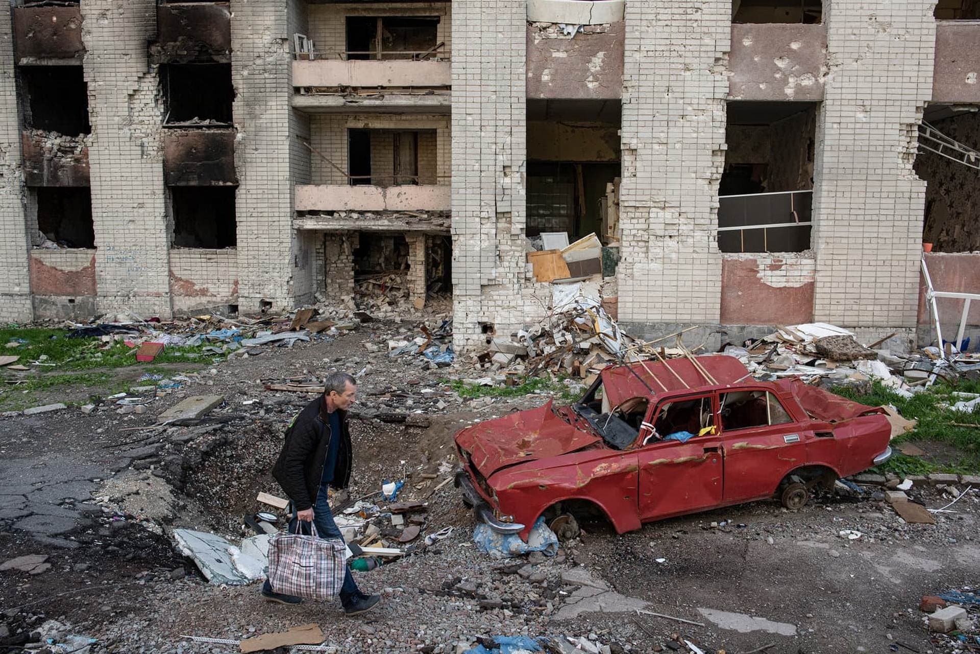 A man carries his belongings past a destroyed car next to a heavily damaged apartment building in Chernihiv
