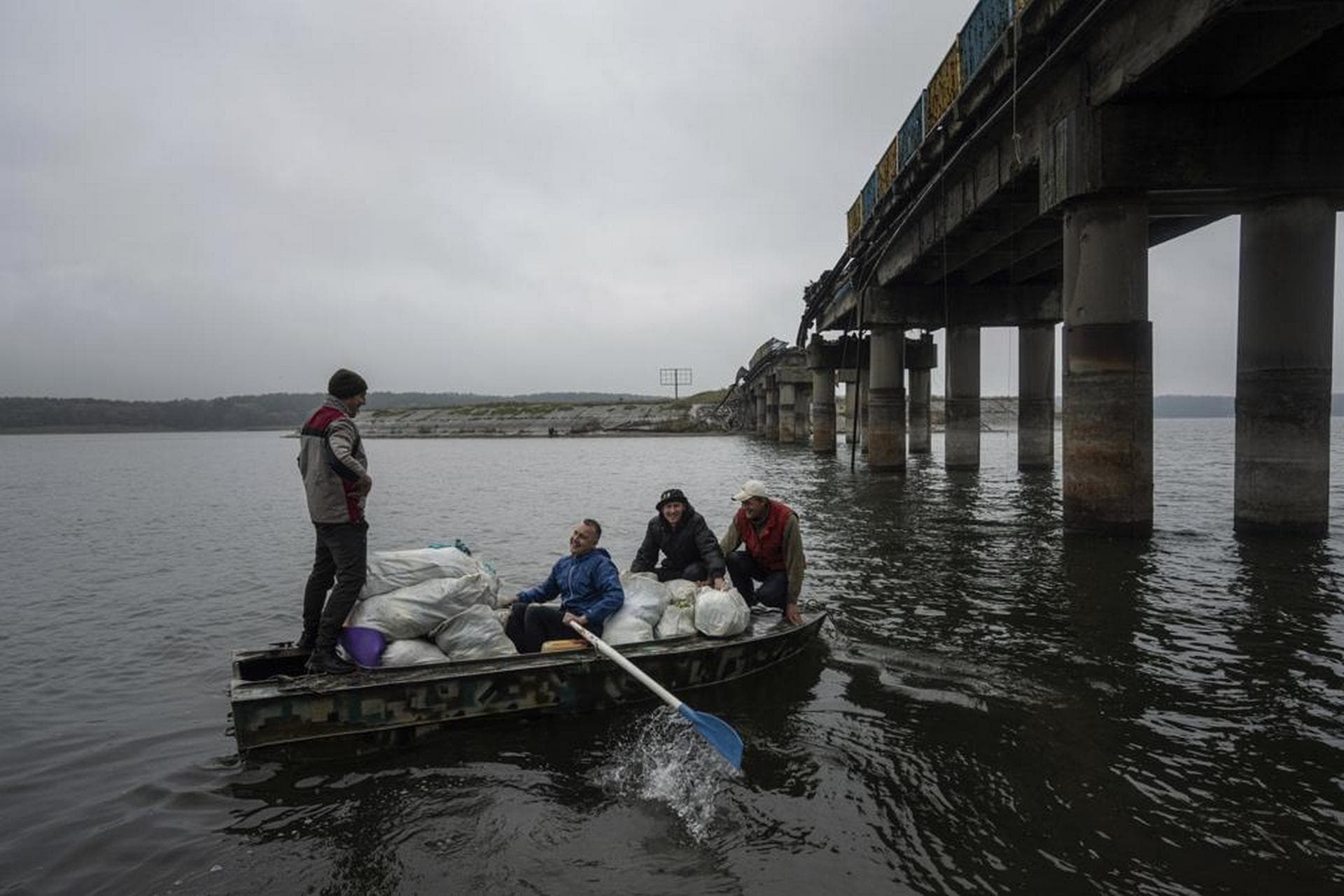Men on a boat transport humanitarian aid across the Siverskyi-Donets river in front of a destroyed bridge in Staryi-Saltiv