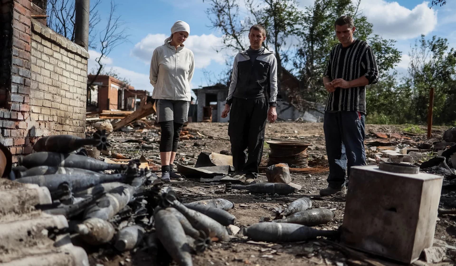 Local residents stand near mortar ammunition at a destroyed street in the village of Kamyanka