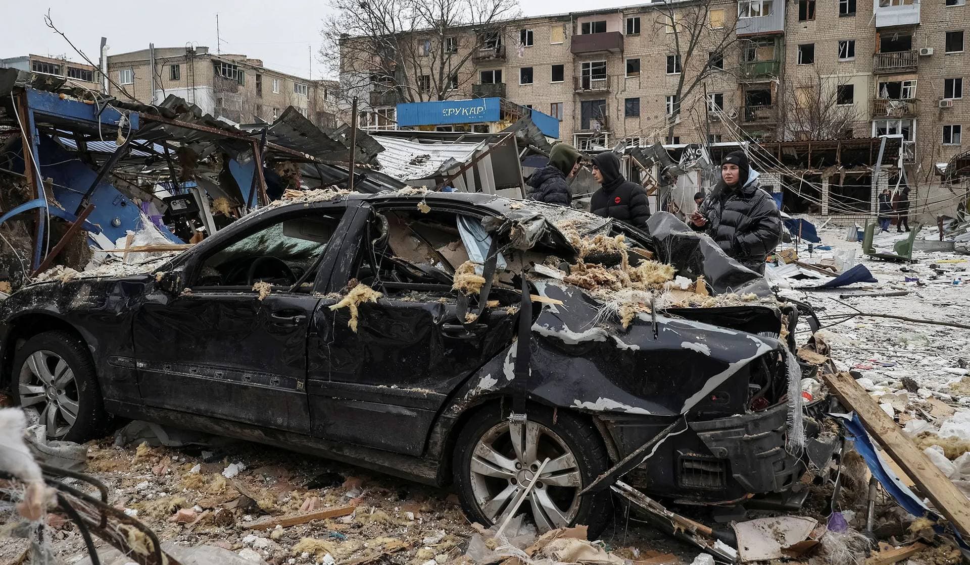 Local residents stand near a destroyed car at a site of apartment building damaged during a Russian missile strike in Kharkiv