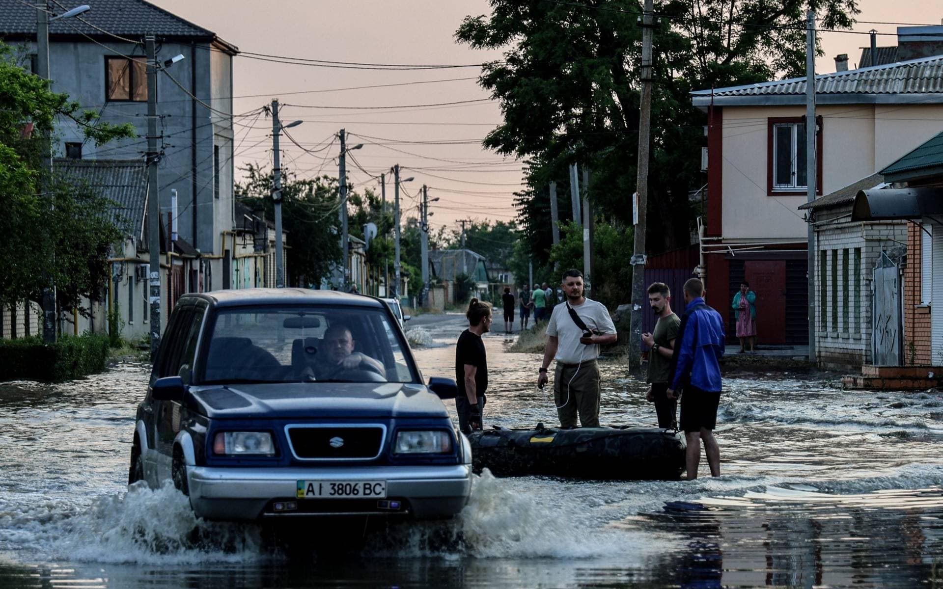 A car makes its way past people standing next to an inflatable boat, in a flooded street of Kherson