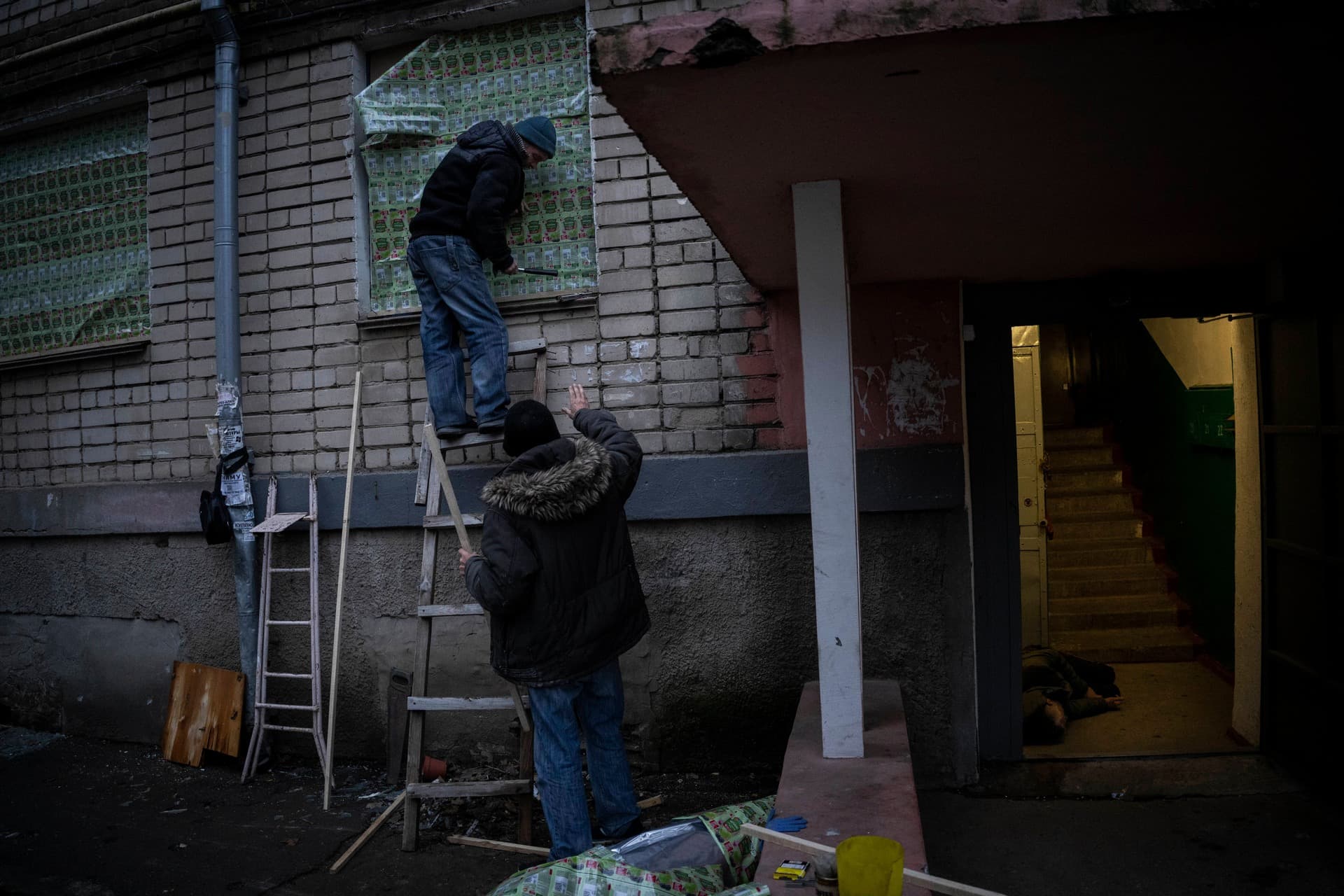 Two residents use food-packaging material to cover windows of a damaged building during a recent Russian strike while another resident lies inebriated at the entrance of a building in Kherson