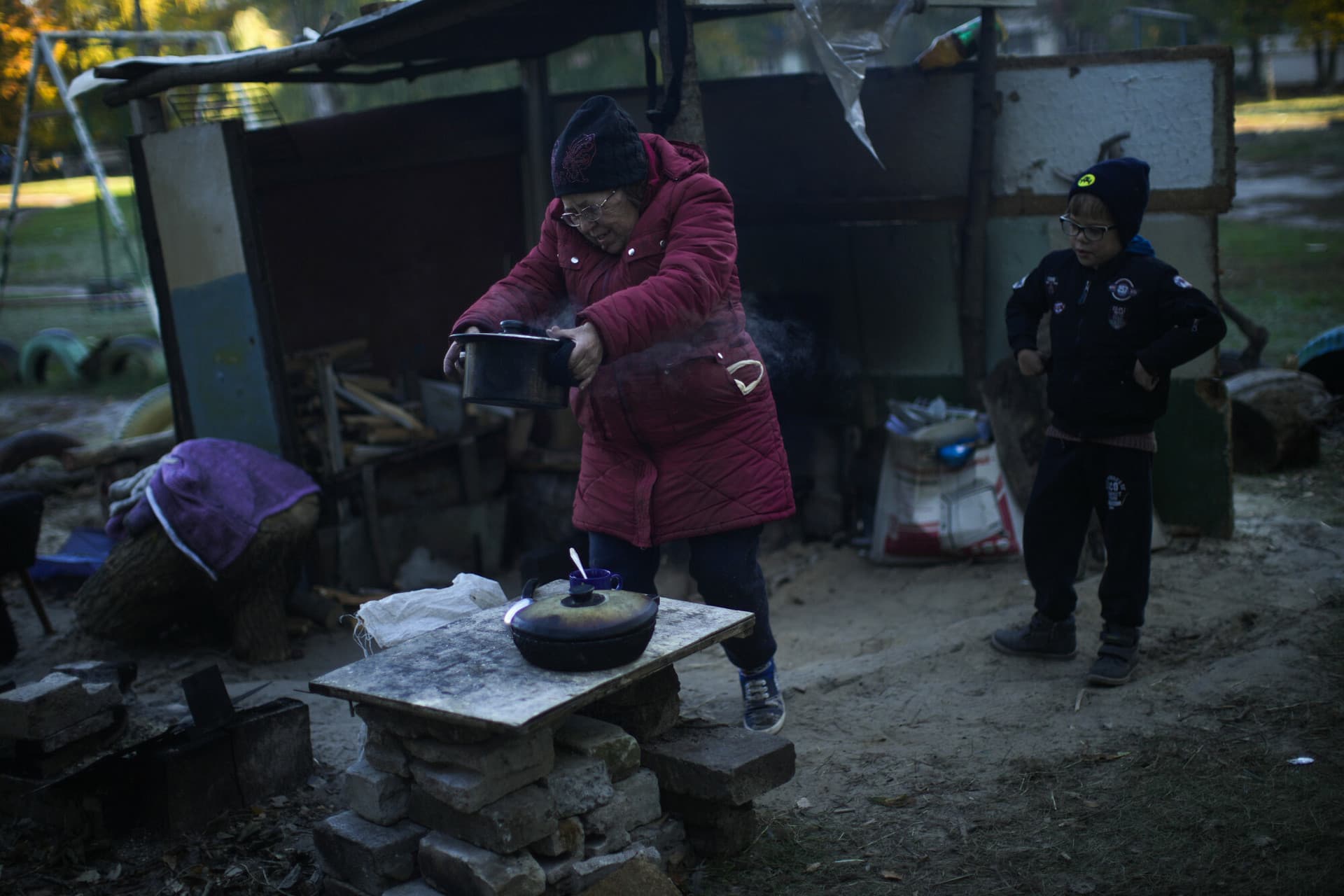 Iryna Panchenko removes a pot of food from a makeshift stove next to her grandchild Artem in Kivsharivka