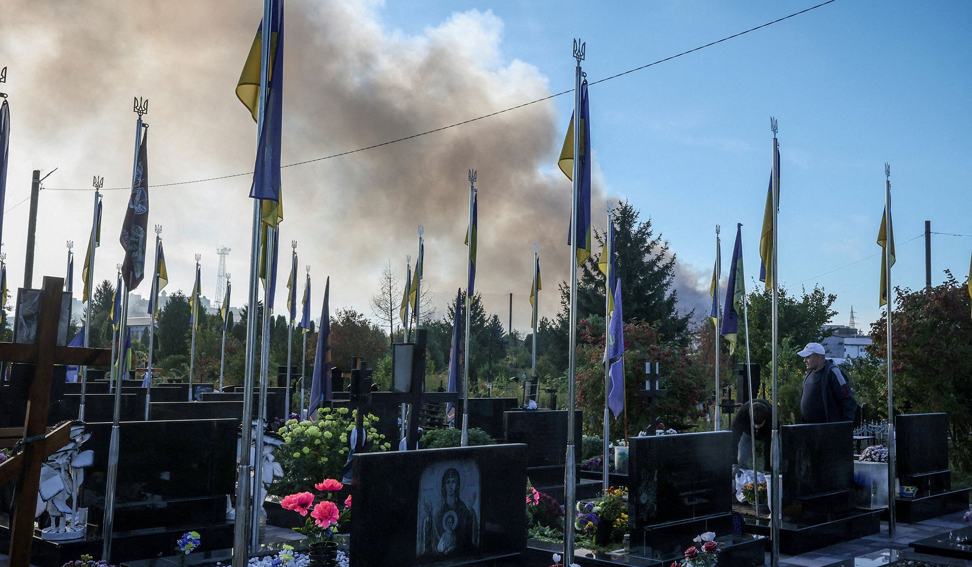 Men clean the graves of fallen soldiers at a local cemetery as smoke rises in the background following a Russian drone strike in Kyiv Region, Ukraine, September 23, 2026.