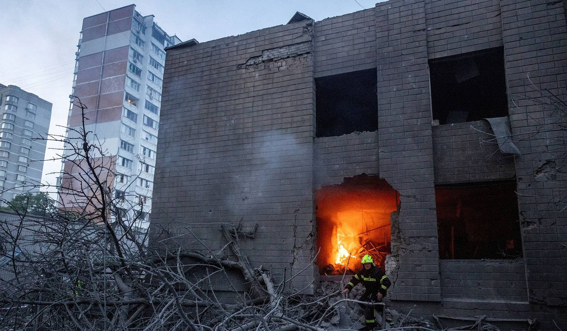 A firefighter stands in front of a burning building next to an apartment building damaged and partially collapsed during Russian missile and drone strikes in Kyiv