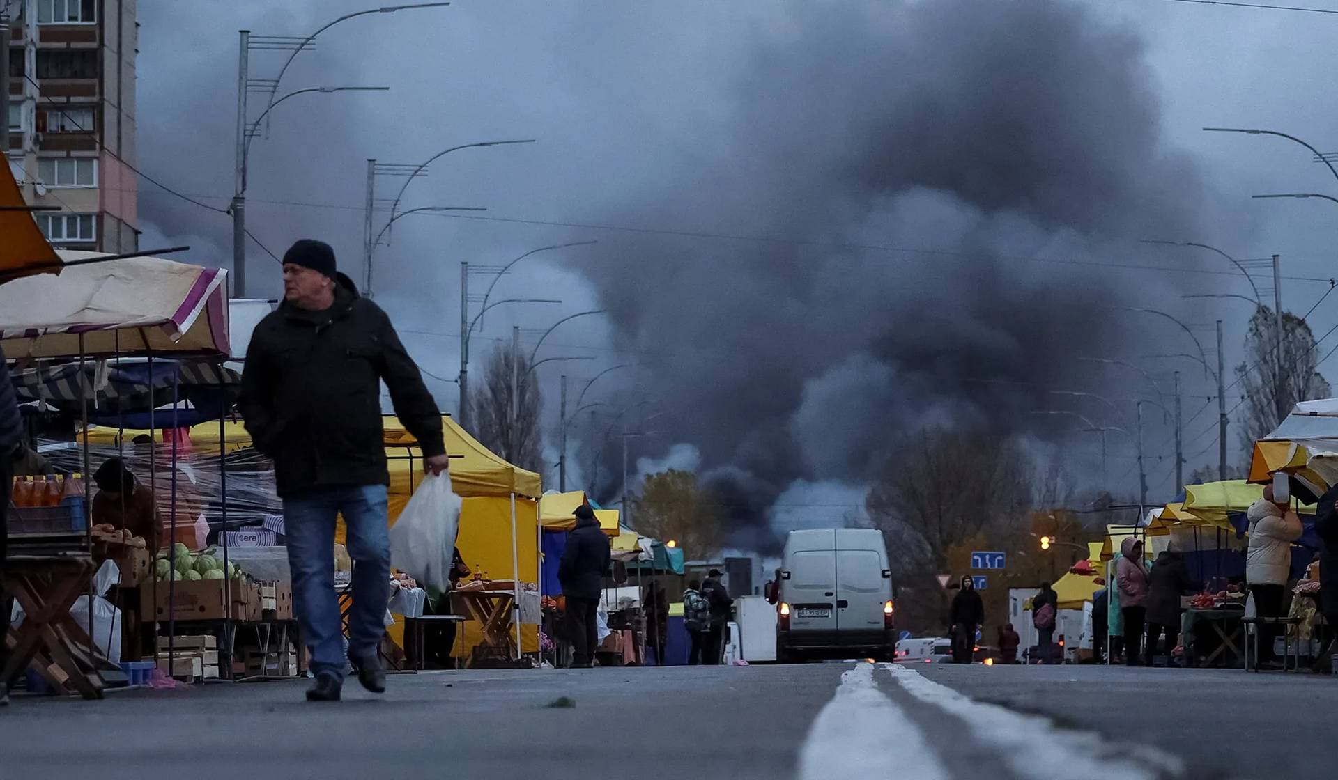 Residents buy groceries at a street market as smoke rises at the site of food warehouses hit by an overnight Russian missile strike in Kyiv