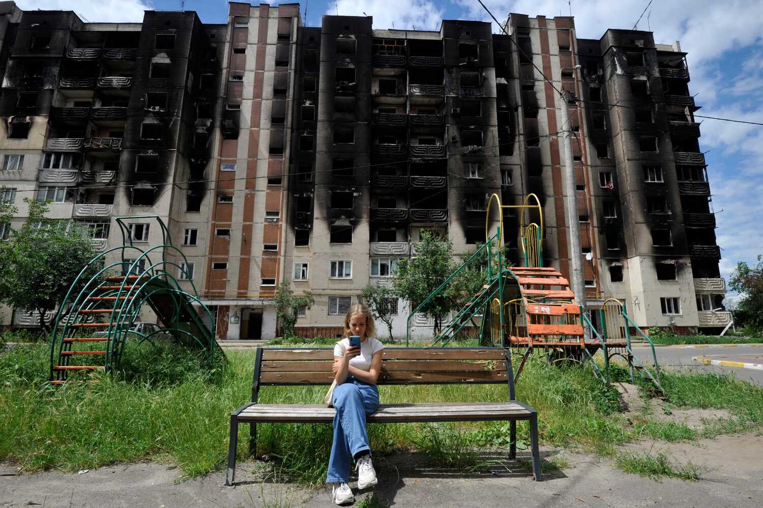 The girl looks at the smartphone on a bench in front of a destroyed multi -storey building in Kyiv