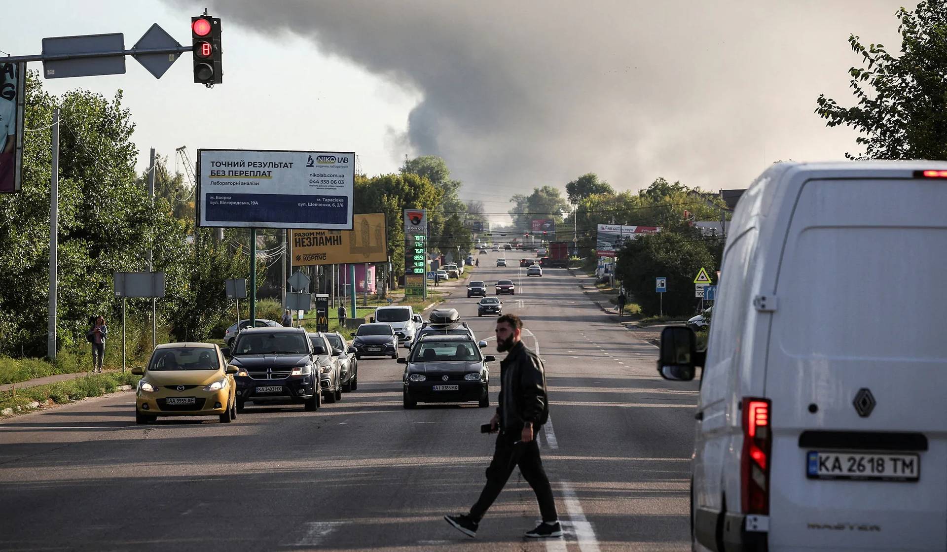 A man crosses a road as smoke rises in the background following an overnight Russian drone strike in Kyiv Region