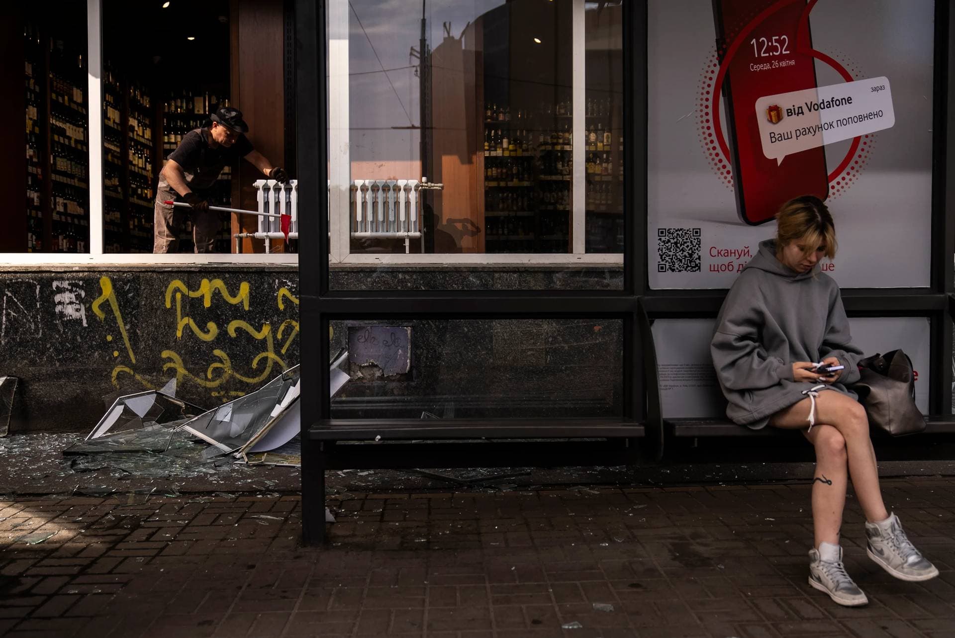A commuter waits for a bus in front of a liquor store damaged in Russian drone attacks in Kyiv