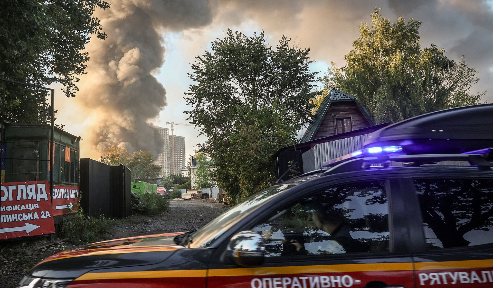 An emergency service vehicle drives by the site of a Russian drone strike as smoke rises in the background in Kyiv