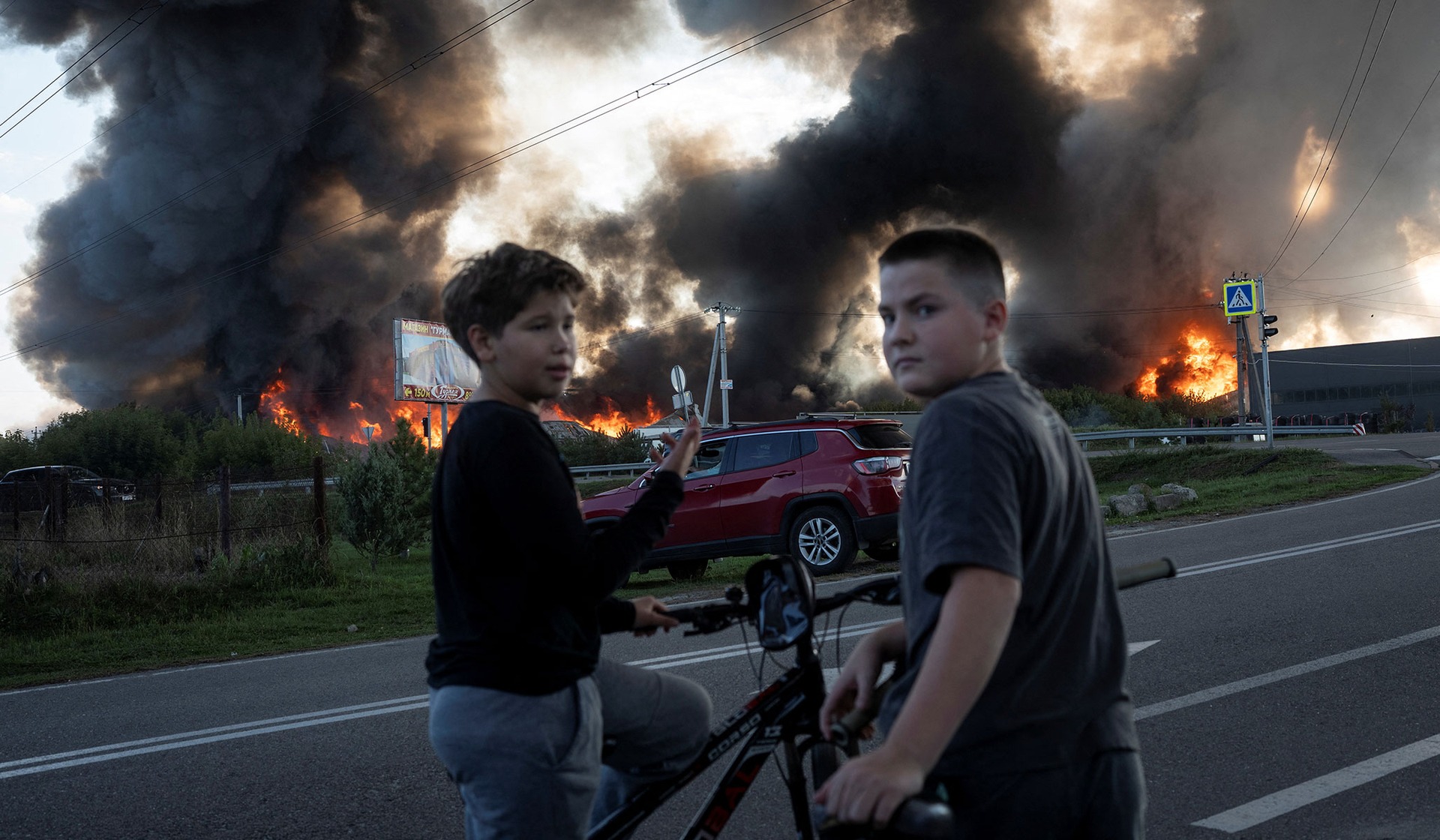 Boys with bicycles stand on the road as smoke rises over a warehouse hit by a Russian drone strike in Kyiv Region, Ukraine, September 20, 2026.