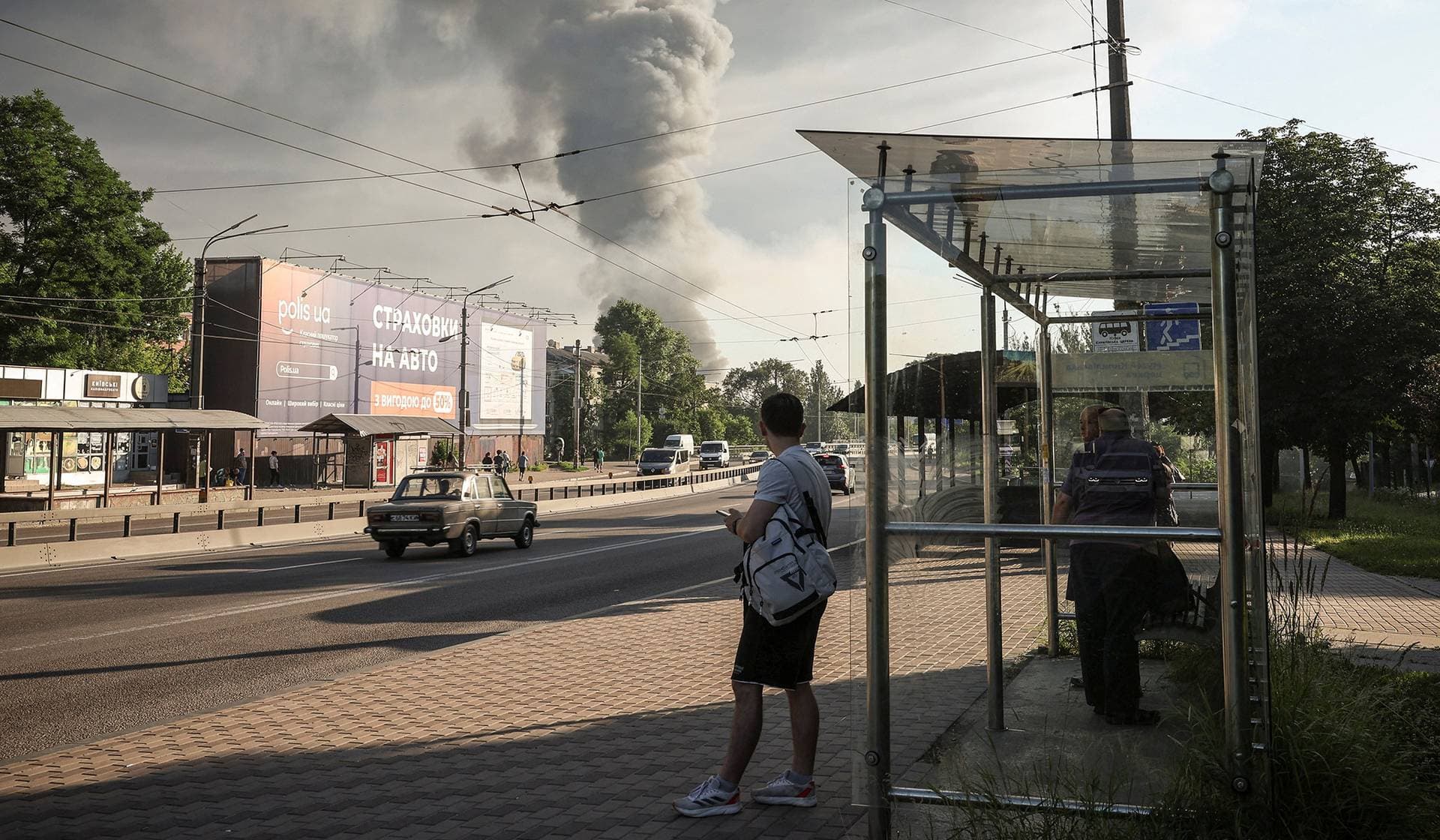People stand at a bus stop as smoke rises following Russian missile and drone strikes in Kyiv