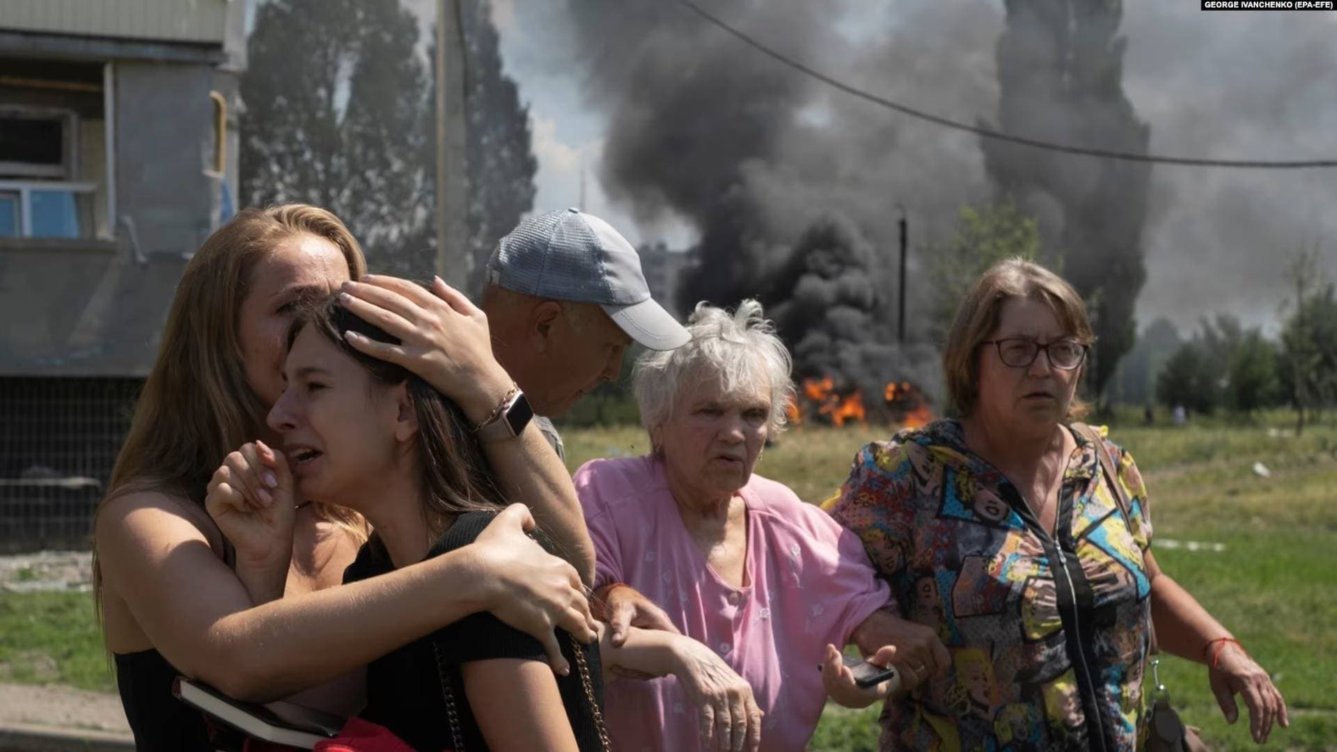 A young girl is comforted by a woman as others help an elderly woman as cars burn in the background