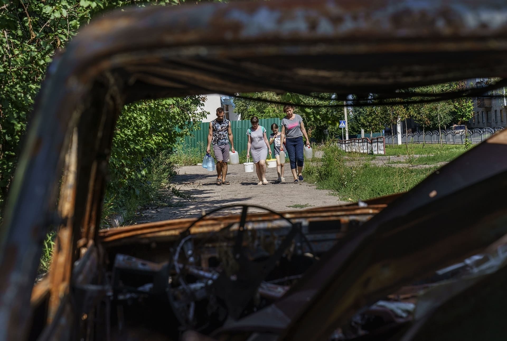 Residents carry water collected at a nearby well while walking past the charred remains of a car from a May rocket attack in Sloviansk
